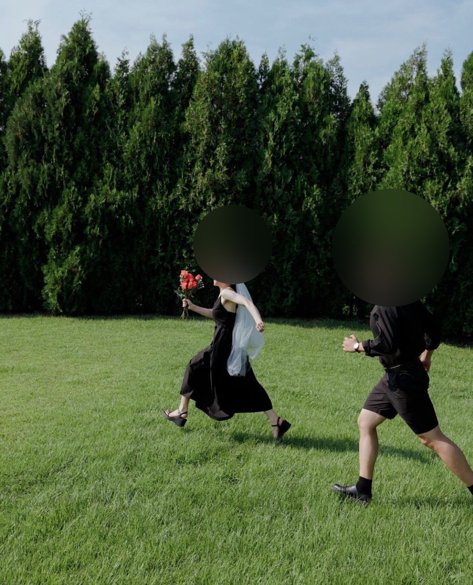 신부는 블랙 끈 원피스에 흰 숏베일, 신랑은 블랙 셔츠와 반바지 차림으로 높은 측백나무 울타리 앞 잔디를 뛰어가는 컷이에요. 두 레퍼런스의 블랙 룩과 잔디·울타리 배경이 한 장에 다 들어 있어요.