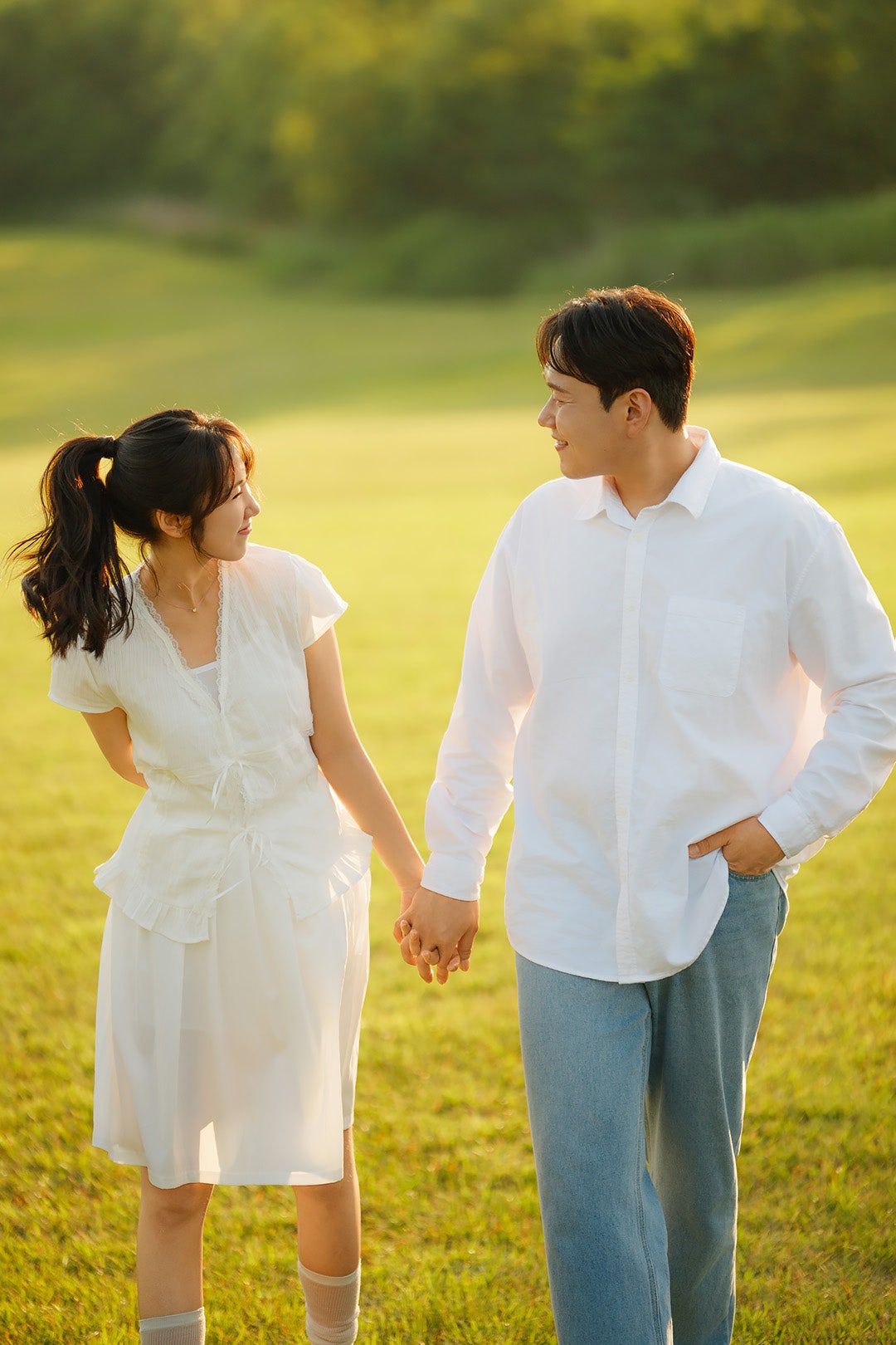 역광 받는 노을공원 잔디에서 손잡고 마주 보며 웃는 컷이에요. 신랑 흰 셔츠와 청바지 조합이 자연스러워요.